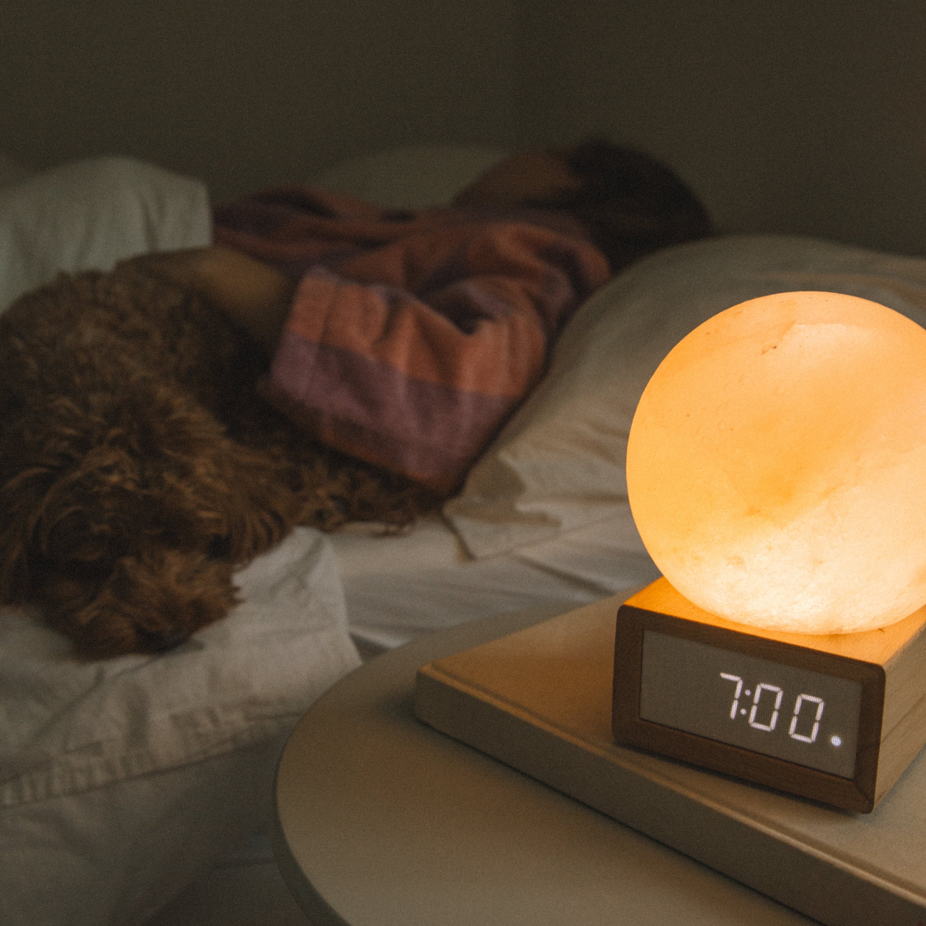 Himalayan salt lamp with digital clock on a bedside table next to a person in bed.